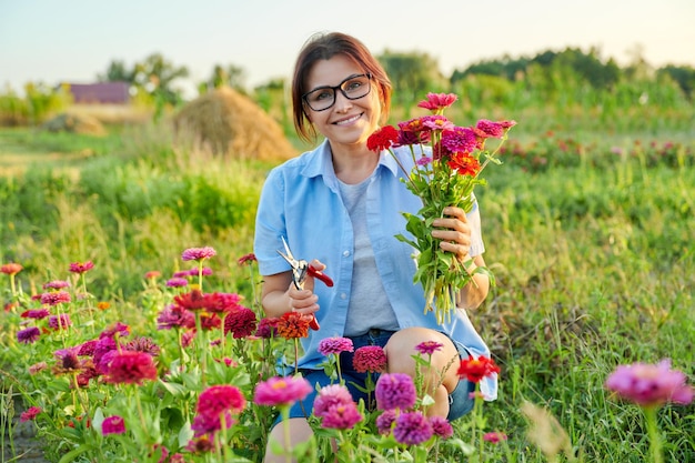 Femme d'âge moyen souriante tenant un bouquet de fleurs de zinnia fraîches dans le jardin