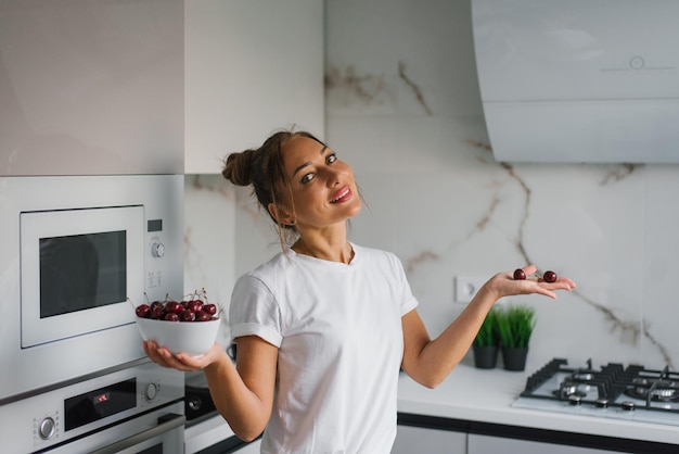 Une femme d'âge moyen avec un sourire se tient dans la cuisine tenant une assiette avec des baies de cerise fraîches et des sourires