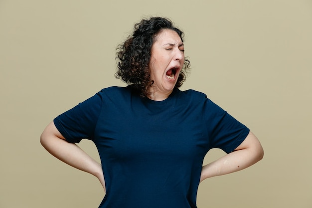 Femme d'âge moyen stressée portant un t-shirt gardant les mains sur la taille criant avec les yeux fermés isolé sur fond vert olive
