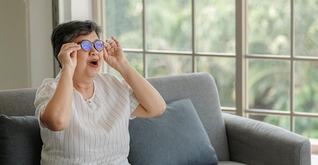 Femme d'âge moyen surprise mettant des lunettes et regardant ailleurs alors qu'elle était assise sur un canapé près de la fenêtre à la maison