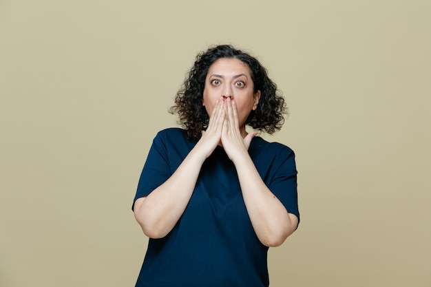 Femme d'âge moyen surprise portant un t-shirt regardant la caméra en gardant les mains sur la bouche faisant un geste oops isolé sur fond vert olive