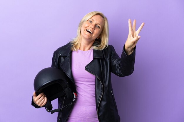 Femme d'âge moyen tenant un casque de moto isolé sur mur violet heureux et compter trois avec les doigts