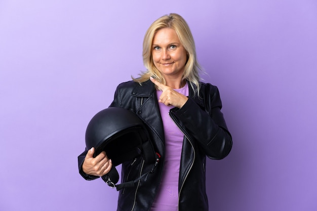 Femme d'âge moyen tenant un casque de moto isolé sur violet pointant vers le côté pour présenter un produit