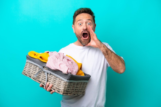 Femme d'âge moyen tenant un panier à vêtements isolé sur fond bleu avec une expression faciale surprise et choquée