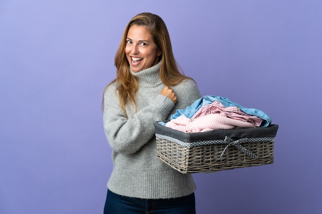 Femme d'âge moyen tenant un panier de vêtements isolé sur mur violet célébrant une victoire