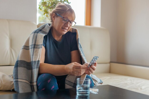 Une femme d'âge moyen tient une plaquette de pilules et lit des instructions médicales assise sur le canapé