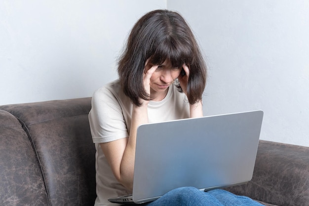 Une femme d'âge moyen triste et frustrée se sent fatiguée de masser ses tempes s'inquiète d'un problème alors qu'elle est assise sur un canapé avec un ordinateur portable Concept d'heures supplémentaires Concept de santé mentale