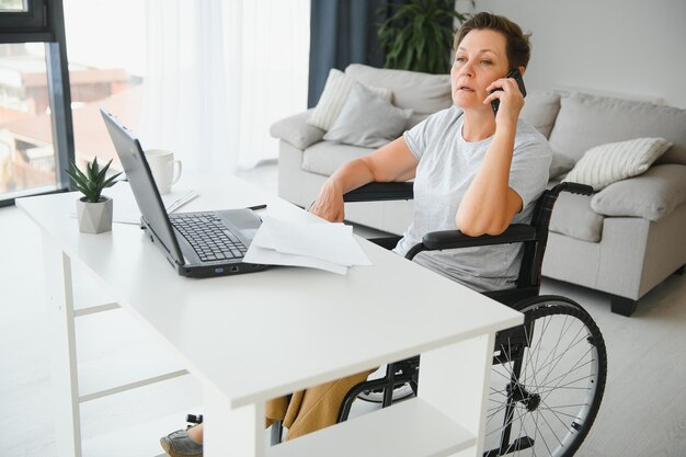 Femme d'âge moyen utilisant un ordinateur portable assis sur un fauteuil roulant à la maison