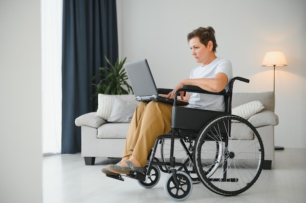 Femme d'âge moyen utilisant un ordinateur portable assis sur un fauteuil roulant à la maison