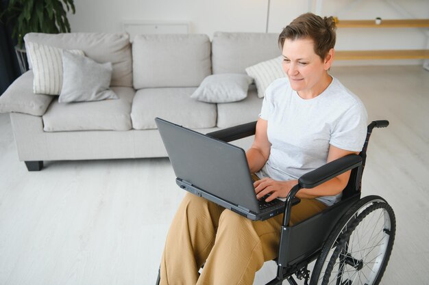 Femme d'âge moyen utilisant un ordinateur portable assis sur un fauteuil roulant à la maison