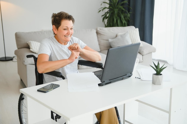 Femme d'âge moyen utilisant un ordinateur portable assis sur un fauteuil roulant à la maison