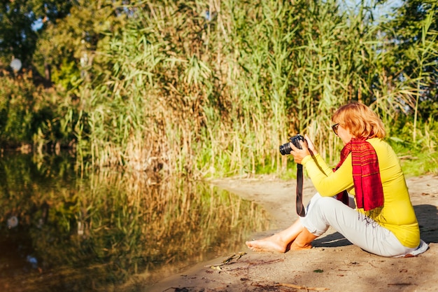 Femme d'âge moyen vérifiant des images sur un appareil photo assis au bord de la rivière en automne. Femme aînée appréciant des photos de prise de vue de nature et de passe-temps