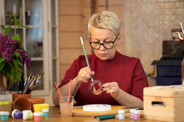 femme d'âge moyen vif peint un bracelet en bois dans une maison de campagne. passe-temps du dessin.