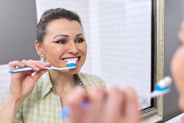 Femme d'âge mûr se brosser les dents dans la salle de bain, se regarder dans le miroir, hygiène dentaire matin et soir