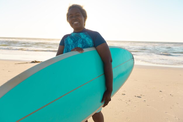 Une femme âgée afro-américaine avec une planche de surf jouissant sur une plage de sable contre la mer et le ciel. Copier l'espace, les sports nautiques, les loisirs, la retraite, inchangée, heureuse, vacances, coucher de soleil, nature.