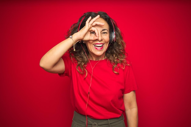 Une femme âgée d'âge moyen portant des écouteurs écoutant de la musique sur un fond rouge isolé faisant un geste ok avec la main souriant un œil regardant à travers les doigts avec un visage heureux