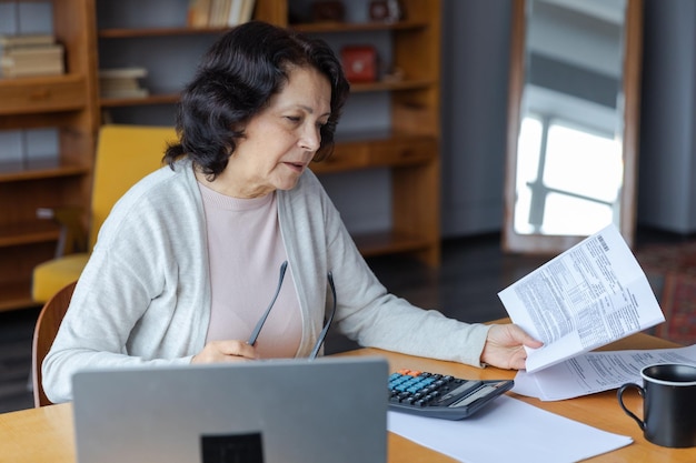 Femme âgée d'âge moyen s'asseoir avec un ordinateur portable et un document papier pensive dame mature plus âgée lisant du papier