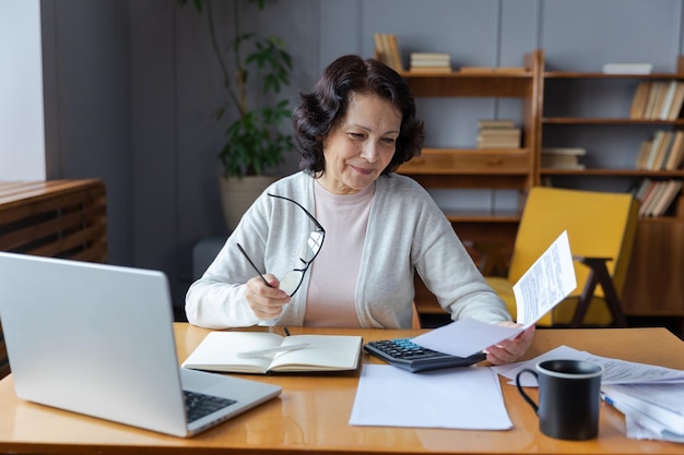 Femme âgée d'âge moyen s'asseoir avec un ordinateur portable et un document papier Sourire d'une dame mûre plus âgée lisant une facture papier payer en ligne à la maison gérer les finances bancaires calculer les impôts planifier le paiement de la pension de la dette de prêt