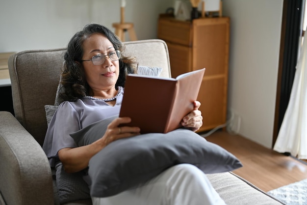Femme âgée asiatique calme portant des lunettes lisant un livre en position allongée sur un canapé