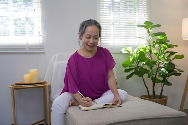 Une femme âgée asiatique détendue et heureuse écrit son idée sur un bloc-notes alors qu'elle est assise sur le canapé de son salon.