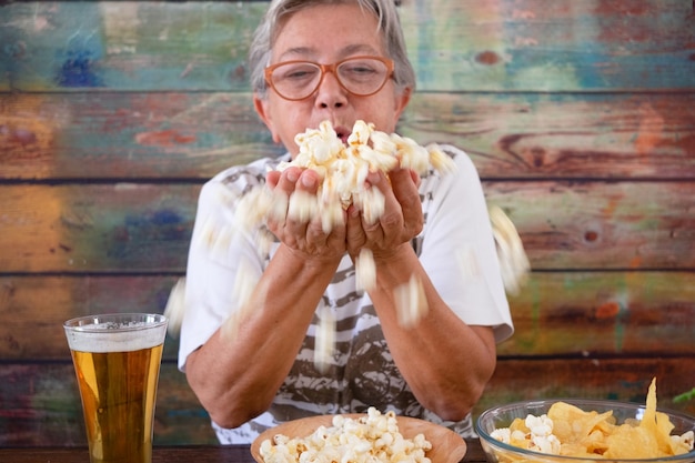 Femme âgée assise à une table en bois avec de la bière et de l'apéritif soufflant sur du pop corn dans ses mains