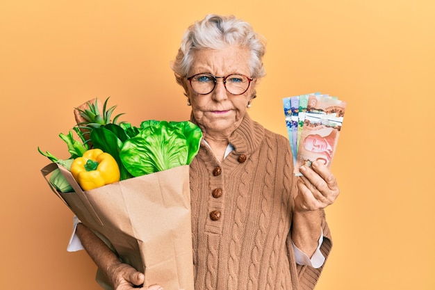 Femme âgée aux cheveux gris tenant des produits d'épicerie et des dollars canadiens sceptique et nerveuse fronçant les sourcils bouleversée à cause d'un problème de personne négative