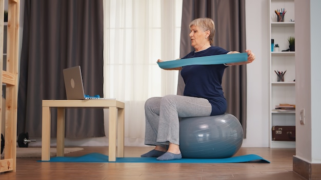 Femme âgée sur ballon de stabilité faisant de l'exercice avec une bande de résistance en regardant une leçon en ligne. Formation en ligne technologie d'apprentissage vieille femme formation de levage mode de vie sain sport fitness entraînement à la maison wi