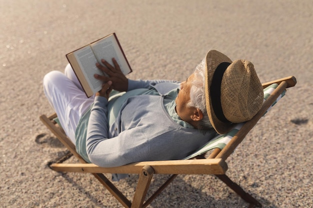 Femme âgée biraciale portant un chapeau lisant un livre tout en se relaxant sur une chaise pliante à la plage