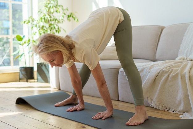Femme âgée en bonne santé active debout dans une pose de yoga pour chien orientée vers le bas tout en pratiquant le yoga à la maison