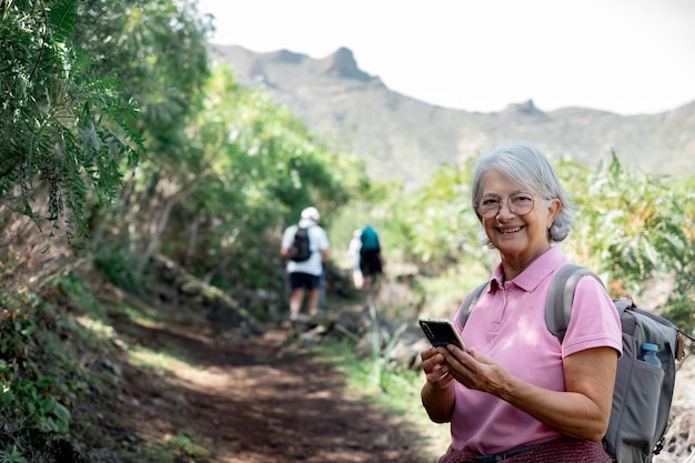 Une femme âgée caucasienne heureuse avec un sac à dos regarde la caméra en parcourant le sentier lors d'une randonnée en montagne