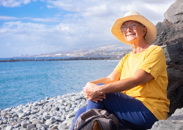 Femme âgée décontractée portant un chapeau de paille assise sur une plage de galets en mer regardant loin