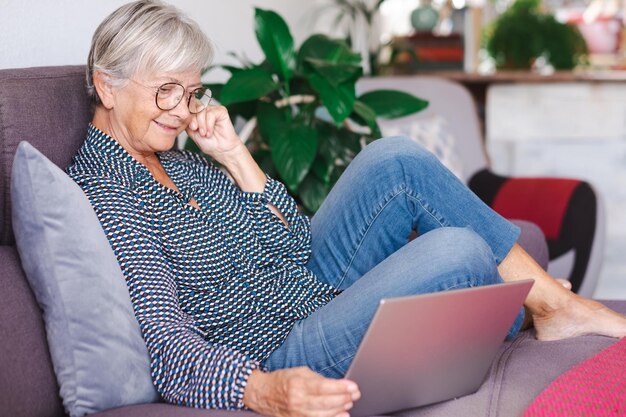 Femme âgée détendue travaillant sur un ordinateur portable à la maison assise sur un canapé dans le salon Dame senior moderne appréciant la technologie et les réseaux sociaux