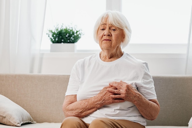 Femme âgée douleur intense au cœur assise sur le canapé problèmes de santé dans la vieillesse vie de mauvaise qualité grand-mère aux cheveux gris tenant sur sa poitrine crise cardiaque et problème de pression d'AVC