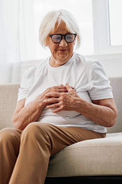 Femme âgée douleur thoracique sévère assise sur le canapé problèmes de santé dans la vieillesse mauvaise qualité de vie grand-mère aux cheveux gris tient sa poitrine avec ses mains santé des femmes cancer du sein