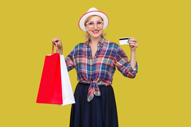 Femme âgée drôle en chapeau blanc et lunettes roses, en chemise à carreaux debout, tenant une carte de crédit bancaire et des sacs à provisions avec un sourire à pleines dents, regardant la caméra. Intérieur, tourné en studio, fond jaune