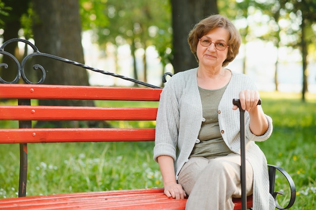 Une femme âgée élégante en chemise est assise sur le banc dans un parc par une chaude journée