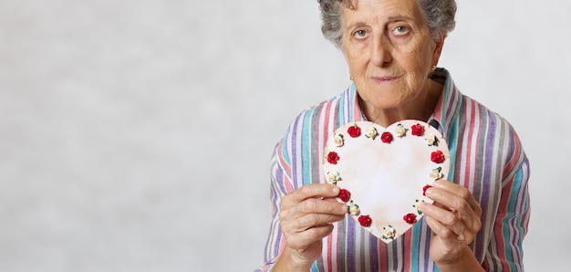 Une femme âgée entre 70 et 80 ans garde le cœur entre les mains. Fond gris