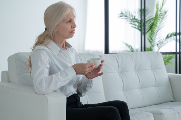 Une femme âgée est assise sur le canapé, boit du café ou du thé et regarde par la fenêtre