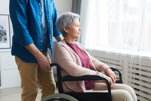 Femme âgée handicapée assise en fauteuil roulant et regardant par la fenêtre avec un soignant debout à proximité