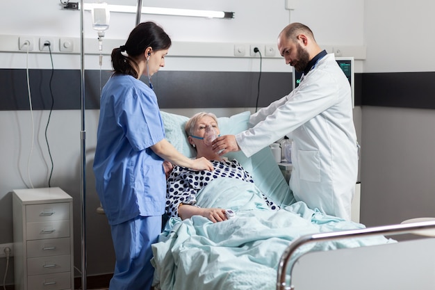 Une femme âgée hospitalisée inspire et expire à travers un masque à oxygène posé dans un lit d'hôpital. Infirmière médicale utilisant le stéthoscope écoutant le coeur patient