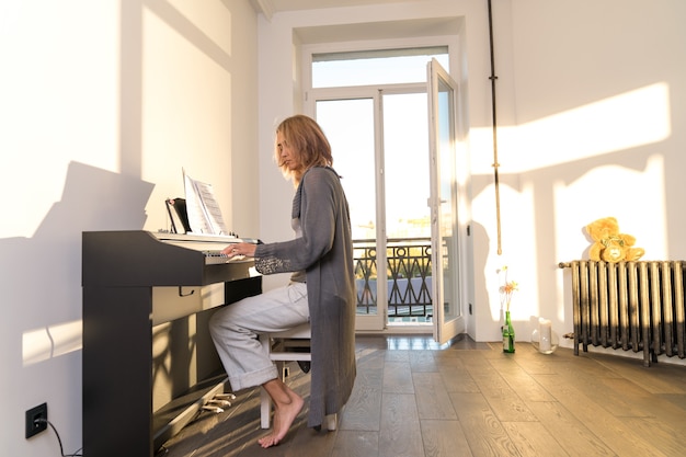 Une femme âgée joue du piano électronique dans une pièce à travers les fenêtres de laquelle bat la lumière du soleil