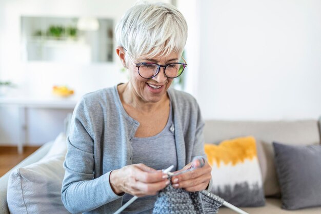 Une femme âgée à lunettes est assise sur un canapé à la maison, souriante tenant des aiguilles à tricoter et du fil tricote des vêtements pour ses proches, son activité préférée et son passe-temps, concept de vie tranquille et insouciant à la retraite