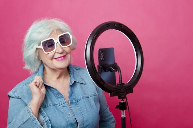 femme âgée avec des lunettes de soleil rétro posant dans une salopette en jean devant un téléphone portable et un anneau lumineux