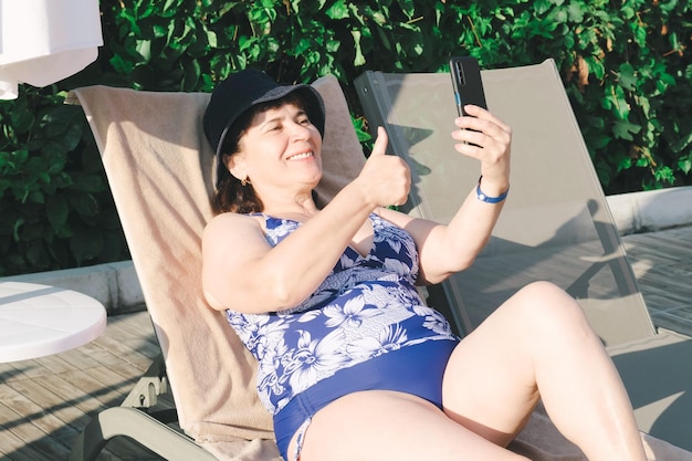 Une femme âgée en maillot de bain sur une chaise longue au bord de la piscine prend un selfie sur un smartphone et montre les pouces vers le haut