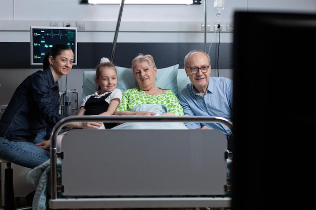 Femme âgée malade allongée dans le lit de la chambre d'hôpital avec équipement de surveillance médicale regardant un programme télévisé sur écran plat dans la chambre de la clinique gériatrique avec des membres proches de la famille.