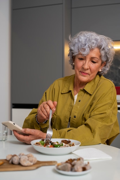 Femme âgée mangeant un plat de figues à la maison et utilisant un smartphone