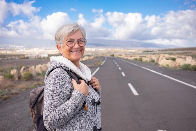 Femme âgée mature attrayante et heureuse marchant seule à l'extérieur sur une route de campagne tenant un sac à dos
