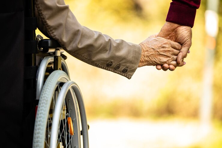 Une femme âgée méconnaissable en fauteuil roulant tenant la main d'un homme âgé.