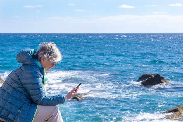 Femme âgée en mer en journée d'hiver, assise sur les rochers en regardant un téléphone intelligent en souriant. Beauté de la nature et horizon au-dessus de l'eau