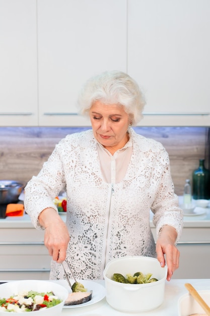 Une femme âgée met du poisson cuit à la vapeur et du brocoli dans une assiette dans la cuisine